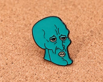 Puede incluir: Un pin de esmalte color cerceta que muestra la cabeza de un personaje con una mandíbula prominente, párpados pesados y labios fruncidos. El diseño está delineado en metal negro, capturando una expresión facial estilizada con rasgos definidos.
