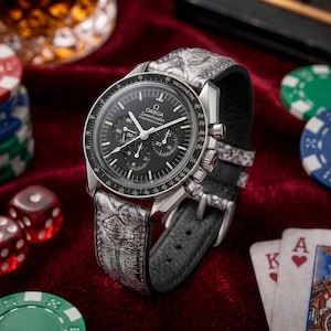 May include: An Omega Speedmaster Professional chronograph watch with a black dial, white hands, and a tachymeter bezel. The watch features a textured, grey, scale-patterned leather strap with a silver-toned buckle and a stainless steel case.