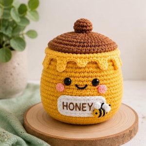 May include: A yellow crocheted honey pot with a brown lid and a white label featuring the text HONEY. The pot has a smiling face with black eyes, pink cheeks, and a small crocheted bee attached to the front label, all crafted in yarn.