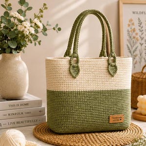 May include: A two-toned crochet tote bag featuring a cream-colored upper section and a sage green lower section. The bag has two matching sage green braided handles attached with leaf-shaped accents and a small rectangular brown leather label.