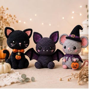 Puede incluir: Tres animales de peluche tejidos a ganchillo que incluyen un gato negro con un lazo naranja y un dije de calabaza, un murciélago morado oscuro con orejas puntiagudas y alas, y un ratón gris con sombrero de bruja negro y capa morada.