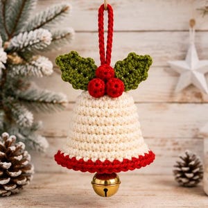 Può includere: Un ornamento a forma di campana realizzato all'uncinetto color crema con un bordo rosso alla base, sormontato da tre bacche rosse e due foglie di agrifoglio verdi, appeso a un cappio di filo rosso con un piccolo campanello in metallo dorato.