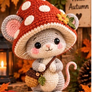 Könnte beinhalten: Eine gehäkelte Mausfigur mit einem großen roten Pilzhut mit weißen Punkten, einer gelben Blume und grünen Blättern. Die Maus hat graues Fell, einen weißen Bauch, rosa Wangen und eine kleine braune Tasche in Eichelform mit einem Blatt.