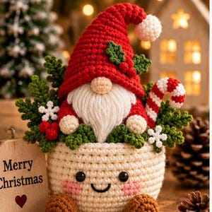 May include: A crocheted gnome with a red hat and white beard sits inside a cream-colored crocheted pot with a smiling face. The gnome is surrounded by green foliage, red berries, a candy cane, and white snowflakes. A wooden sign reads Merry Christmas.