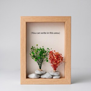 May include: A rectangular light wood shadow box frame containing two small dried flower arrangements, one green and one orange, each secured to a cluster of smooth gray stones. The off-white backing features the text, (You can write in this area.)