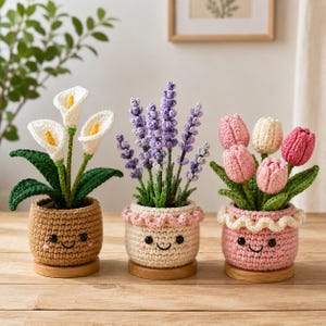 Puede incluir: Tres plantas de ganchillo en maceta con calas blancas, lavanda morada y tulipanes rosas y blancos. Cada planta está en una maceta de ganchillo texturizada con una cara sonriente bordada, sobre una base circular de madera.