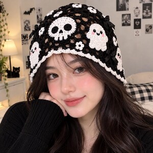 May include: A black and white crocheted beanie featuring a skull motif, ghost figures, and small white flowers. The hat is constructed with an open-weave mesh pattern and is accented with small black bows along the edges and a white scalloped border.