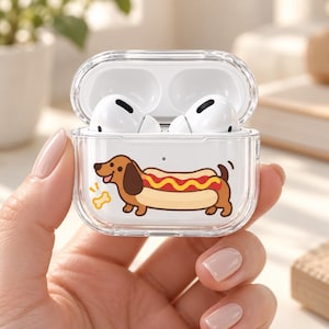 May include: A clear protective case for wireless earbuds featuring a cartoon illustration of a brown dachshund dog shaped like a hot dog with yellow mustard, accompanied by a small yellow bone icon, housing two white wireless earbuds inside.