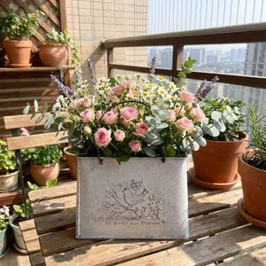 Könnte beinhalten: Ein rechteckiger Metallübertopf mit abgenutzter weißer Oberfläche, verziert mit einem geprägten Vogel auf einem Zweig und dem Text Le Jardin aux Oiseaux. Er ist mit einem Blumenarrangement aus rosa Rosen, weißen Gänseblümchen, Lavendel und Eukalyptus gefüllt.