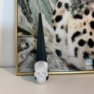 May include: A black cone-shaped ring display stand featuring a translucent white crystal skull ornament attached to the front. The stand has a textured surface and is positioned vertically, showcasing the detailed facial features of the skull.