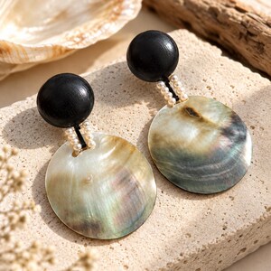 May include: A pair of drop earrings featuring a smooth black wooden sphere at the top, connected by a short strand of small white seed beads to a large, circular, iridescent abalone shell disc with natural swirling patterns of green and gray.