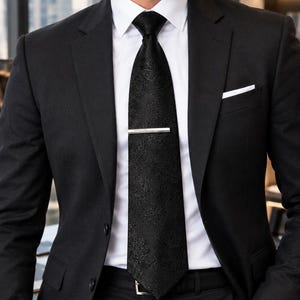 May include: A black suit jacket worn over a crisp white dress shirt with a black paisley patterned necktie. A silver tie bar is fastened horizontally across the centre of the tie. A white pocket square is tucked into the left chest pocket.