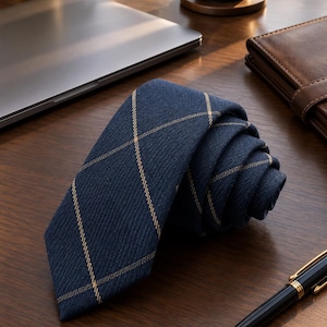 Pode incluir: Uma gravata azul-marinho com um padrão subtil de grelha diagonal em tom castanho-claro. A gravata está enrolada, exibindo o seu tecido texturizado e a ponta. O design consiste em linhas finas que se cruzam formando um motivo de losangos.