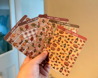 May include: Five rectangular cards labeled WASHI TAPES, each displaying multiple strips of decorative adhesive tape. The designs feature autumn-themed patterns including brown leaves, plaid, small flowers, pies, lanterns, and woodland creatures in warm orange, brown, and cream tones.