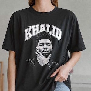 May include: A black short-sleeve t-shirt featuring a white graphic print of the singer Khalid. The design includes the name KHALID in a bold, arched, distressed-style font above a black and white portrait of the artist resting his chin on his hand.