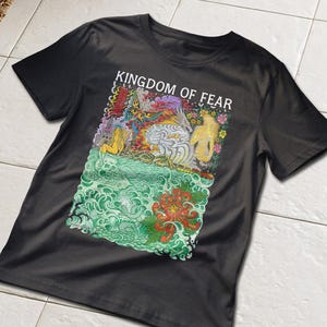 May include: A black short-sleeve t-shirt featuring a colorful graphic print with the text KINGDOM OF FEAR. The design includes a dragon, a skull, a large red flower, swirling green waves, and two figures with long hair amidst a vibrant floral background.