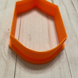 May include: Orange plastic cookie cutter shaped like a shield with a rounded bottom.