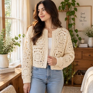 May include: A cream-colored, open-front cardigan featuring a detailed crochet granny square pattern throughout the body and sleeves, with ribbed cuffs and a round neckline, worn over a simple white top and light blue denim jeans.