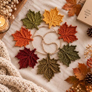May include: A circular garland featuring six knitted maple leaves in shades of olive green, mustard yellow, burnt orange, and deep burgundy, connected by a thin, natural-coloured twisted twine cord, arranged in a circular pattern on a fabric surface.