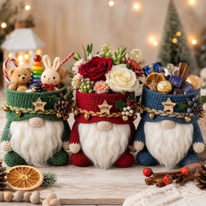Puede incluir: Tres cestas de ganchillo con forma de gnomo en verde, rojo y azul. La cesta verde contiene un osito de peluche, la roja rosas y flores, y la azul chocolates. Cada una tiene una barba blanca, una etiqueta de estrella de madera con la inscripción JOY, LOVE o THANKFUL, y piñas decorativas.