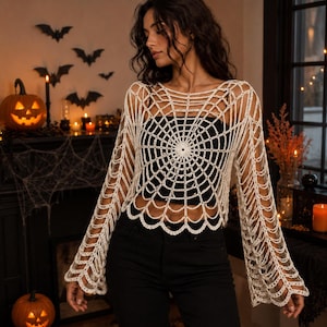 Puede incluir: Una blusa de ganchillo de color crema de manga larga con un intrincado patrón de telaraña radiante que se extiende desde el centro por el pecho y las mangas, terminada con un dobladillo festoneado y puños acampanados.