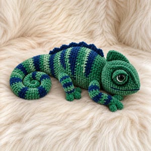 May include: A crocheted chameleon toy featuring a striped pattern in shades of dark blue, forest green, and light green. The plush figure has a curled tail, a textured dorsal crest along its back, and a single large black plastic eye on its side.