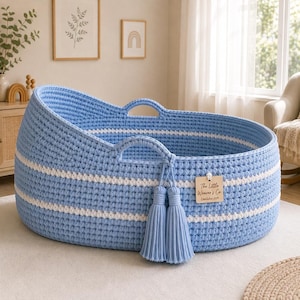 Peut inclure: Un panier ovale tissé bleu clair avec deux poignées intégrées et deux bandes horizontales blanches. Une paire de pompons bleu clair assortis pend à une poignée, et une étiquette en bois indique The Little Weaver's Co. Established 2024.