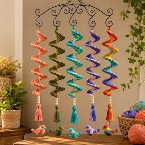 Könnte beinhalten: Fünf hängende Spiral-Häkelornamente an einem schwarzen Metallbügel. Jede Spirale hat einen einzigartigen Farbverlauf in Pink, Grün, Blau, Lila oder Regenbogenfarben, verziert mit weißen Perlen, einer Quaste und einem kleinen gehäkelten Vogel.