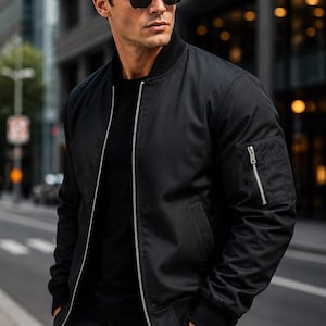 May include: A black bomber jacket featuring a silver front zipper, a ribbed collar, and a zippered utility pocket on the left sleeve. The jacket is worn over a plain black t-shirt and paired with dark trousers, complemented by black sunglasses.