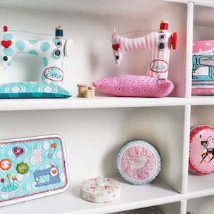 Puede incluir: Una estanter&iacute;a blanca exhibe art&iacute;culos con tem&aacute;tica de costura. Dos m&aacute;quinas de coser de tela con detalles de coraz&oacute;n se encuentran sobre cojines de alfiler a juego. Tambi&eacute;n se ve una bolsa rosa con lunares con un parche de m&aacute;quina de coser, una lata con iconos de costura.