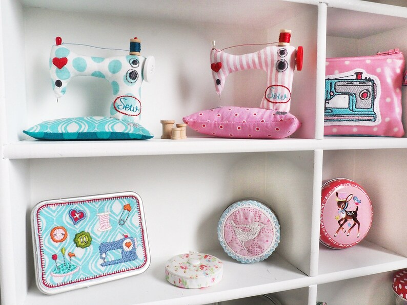 Puede incluir: Una estanter&iacute;a blanca exhibe art&iacute;culos con tem&aacute;tica de costura. Dos m&aacute;quinas de coser de tela con detalles de coraz&oacute;n se encuentran sobre cojines de alfiler a juego. Tambi&eacute;n se ve una bolsa rosa con lunares con un parche de m&aacute;quina de coser, una lata con iconos de costura.