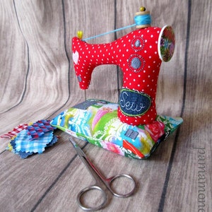 Puede incluir: Una m&aacute;quina de coser de juguete roja con lunares blancos y un carrete de hilo azul descansa sobre un coj&iacute;n floral. Unas tijeras peque&ntilde;as y retales de tela est&aacute;n cerca. La palabra "Sew" est&aacute; bordada en un parche de mezclilla.