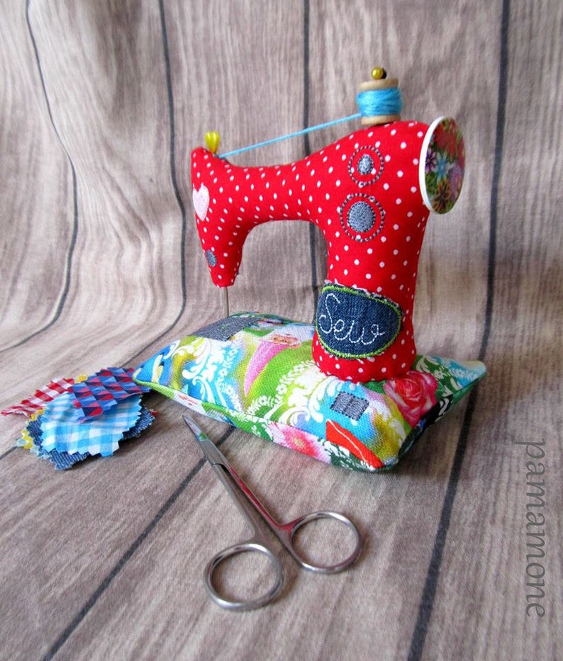 Puede incluir: Una m&aacute;quina de coser de juguete roja con lunares blancos y un carrete de hilo azul descansa sobre un coj&iacute;n floral. Unas tijeras peque&ntilde;as y retales de tela est&aacute;n cerca. La palabra "Sew" est&aacute; bordada en un parche de mezclilla.