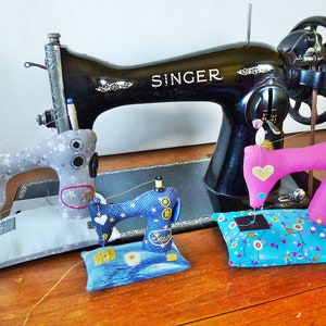 Puede incluir: Una m&aacute;quina de coser Singer negra vintage se encuentra detr&aacute;s de tres m&aacute;quinas de coser de juguete de colores. Los juguetes son grises, azules y rosas, cada uno con un dise&ntilde;o decorativo. La rosa tiene la palabra "Sew".