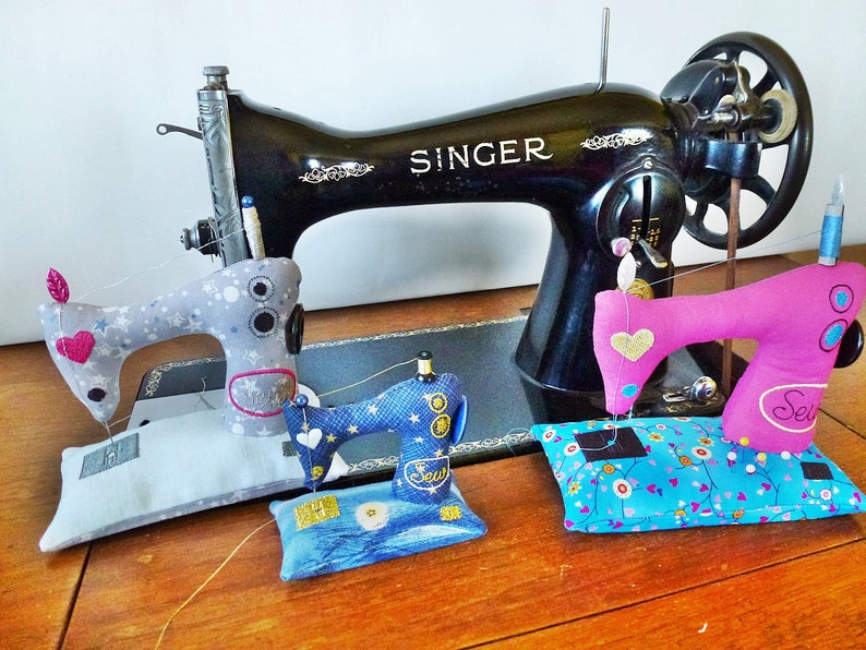 Puede incluir: Una m&aacute;quina de coser Singer negra vintage se encuentra detr&aacute;s de tres m&aacute;quinas de coser de juguete de colores. Los juguetes son grises, azules y rosas, cada uno con un dise&ntilde;o decorativo. La rosa tiene la palabra "Sew".