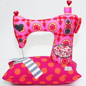 Puede incluir: Una m&aacute;quina de coser de tela rosa caprichosa con un detalle en forma de coraz&oacute;n y una bobina de hilo rosa. La m&aacute;quina descansa sobre una almohada rosa con lunares y una pieza de tela a rayas. La m&aacute;quina tiene un estampado floral y la palabra "Sweet" en el lateral.