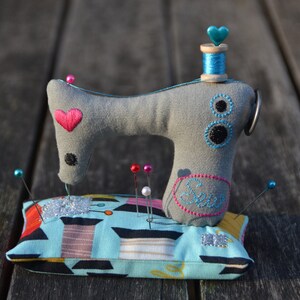 Puede incluir: Un coj&iacute;n para alfileres con forma de m&aacute;quina de coser de tela gris con un coraz&oacute;n rosa y una bobina de hilo azul. El coj&iacute;n tiene un estampado geom&eacute;trico azul y turquesa con alfileres de varios colores. Un accesorio de costura decorativo.