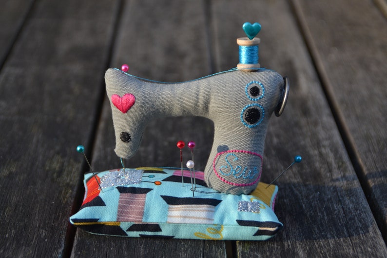 Puede incluir: Un coj&iacute;n para alfileres con forma de m&aacute;quina de coser de tela gris con un coraz&oacute;n rosa y una bobina de hilo azul. El coj&iacute;n tiene un estampado geom&eacute;trico azul y turquesa con alfileres de varios colores. Un accesorio de costura decorativo.