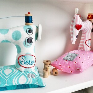 May include: Two miniature sewing machines made of fabric. One is blue and white with polka dots and the word "Sew" embroidered on it. The other is pink and white striped with a red heart. Both are sitting on a white shelf with spools of thread.