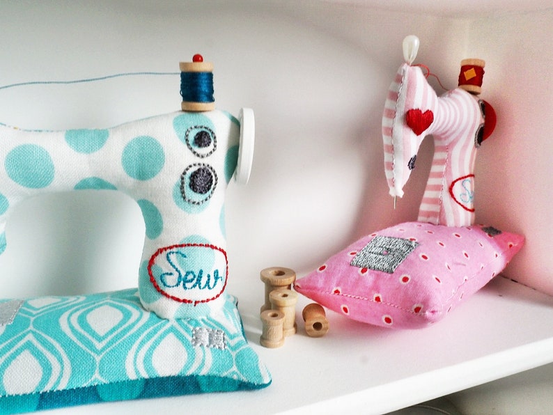 May include: Two miniature sewing machines made of fabric. One is blue and white with polka dots and the word "Sew" embroidered on it. The other is pink and white striped with a red heart. Both are sitting on a white shelf with spools of thread.