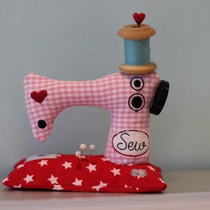 Puede incluir: Una caprichosa figura de m&aacute;quina de coser hecha a mano. El cuerpo de cuadros rosas y blancos presenta detalles de coraz&oacute;n rojo y bot&oacute;n negro. Una bobina de madera con hilo azul y una tapa de coraz&oacute;n rojo se encuentra encima. La base es un coj&iacute;n rojo con estrellas.