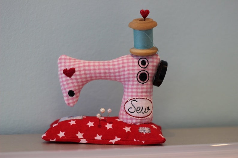 Puede incluir: Una caprichosa figura de m&aacute;quina de coser hecha a mano. El cuerpo de cuadros rosas y blancos presenta detalles de coraz&oacute;n rojo y bot&oacute;n negro. Una bobina de madera con hilo azul y una tapa de coraz&oacute;n rojo se encuentra encima. La base es un coj&iacute;n rojo con estrellas.