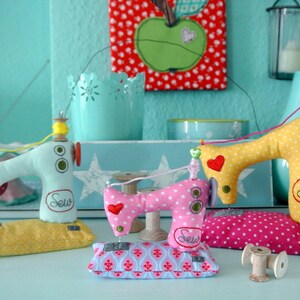 Puede incluir: Tres adornos decorativos de m&aacute;quinas de coser hechos a mano en colores pastel. Las m&aacute;quinas son azul claro, rosa y amarillo, con lunares y detalles de coraz&oacute;n. Cada una tiene un carrete de hilo y la palabra "Sew" bordada en la parte delantera. Se apoyan en cojines decorativos.