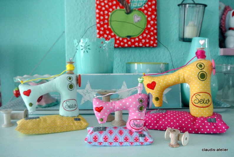 Puede incluir: Tres adornos decorativos de m&aacute;quinas de coser hechos a mano en colores pastel. Las m&aacute;quinas son azul claro, rosa y amarillo, con lunares y detalles de coraz&oacute;n. Cada una tiene un carrete de hilo y la palabra "Sew" bordada en la parte delantera. Se apoyan en cojines decorativos.