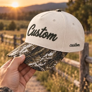 May include: A cream-colored baseball cap featuring a dark green embroidered script word Custom on the front panel and a smaller matching text on the side. The brim displays a dark green and white camouflage pattern with visible stitching.