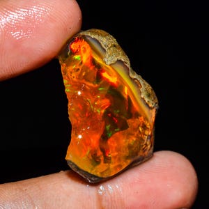 Op de afbeelding: Een ruwe, ongepolijste opaal edelsteen met een levendig kleurenspel met intense oranje, rode en groene flitsen door het doorschijnende lichaam. De steen heeft een onregelmatige, grillige vorm met een ruwe, aardebruine korst aan de bovenrand.