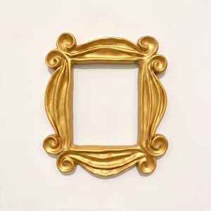 Op de afbeelding: Een rechthoekige fotolijst met een metallic gouden afwerking. De lijst heeft een opvallend, sierlijk ontwerp met prominente, krullende ornamenten op elk van de vier hoeken en golvende, stofachtige texturen langs de randen.