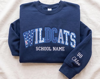 May include: A navy blue crewneck sweatshirt featuring the word WILDCATS in large, patterned applique letters with floral, star, and striped designs. Below is the text SCHOOL NAME in white, and the sleeve displays the number 98 and the name Chloe.