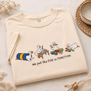 May include: A cream-colored t-shirt featuring a graphic of four white ducks engaged in various activities, including playing in a tunnel, painting at a table, eating from bowls, and doing a puzzle. The text reads, We put the FUN in FUNCTION.