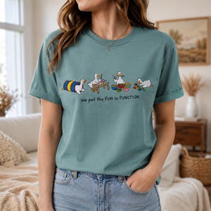 May include: A dusty blue t-shirt featuring a graphic of four white ducks engaged in various activities, including crawling through a tunnel, crafting at a table, eating, and playing with blocks, with the text "We put the FUN in FUNCTION" below.